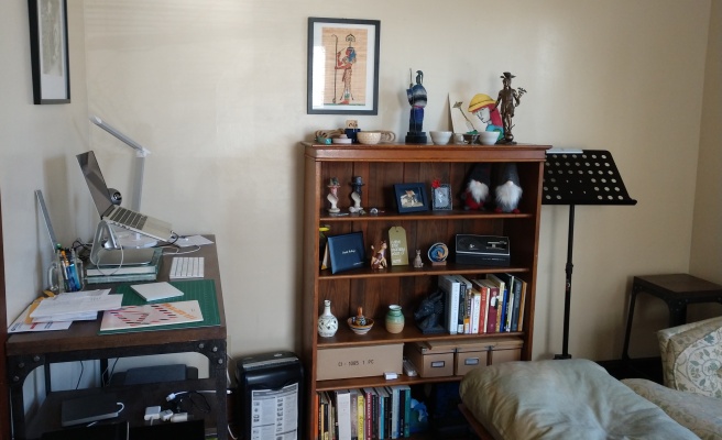 A bookshelf with shrines atop it, with a desk on the left against a perpendicular wall and some soft seating in the foreground